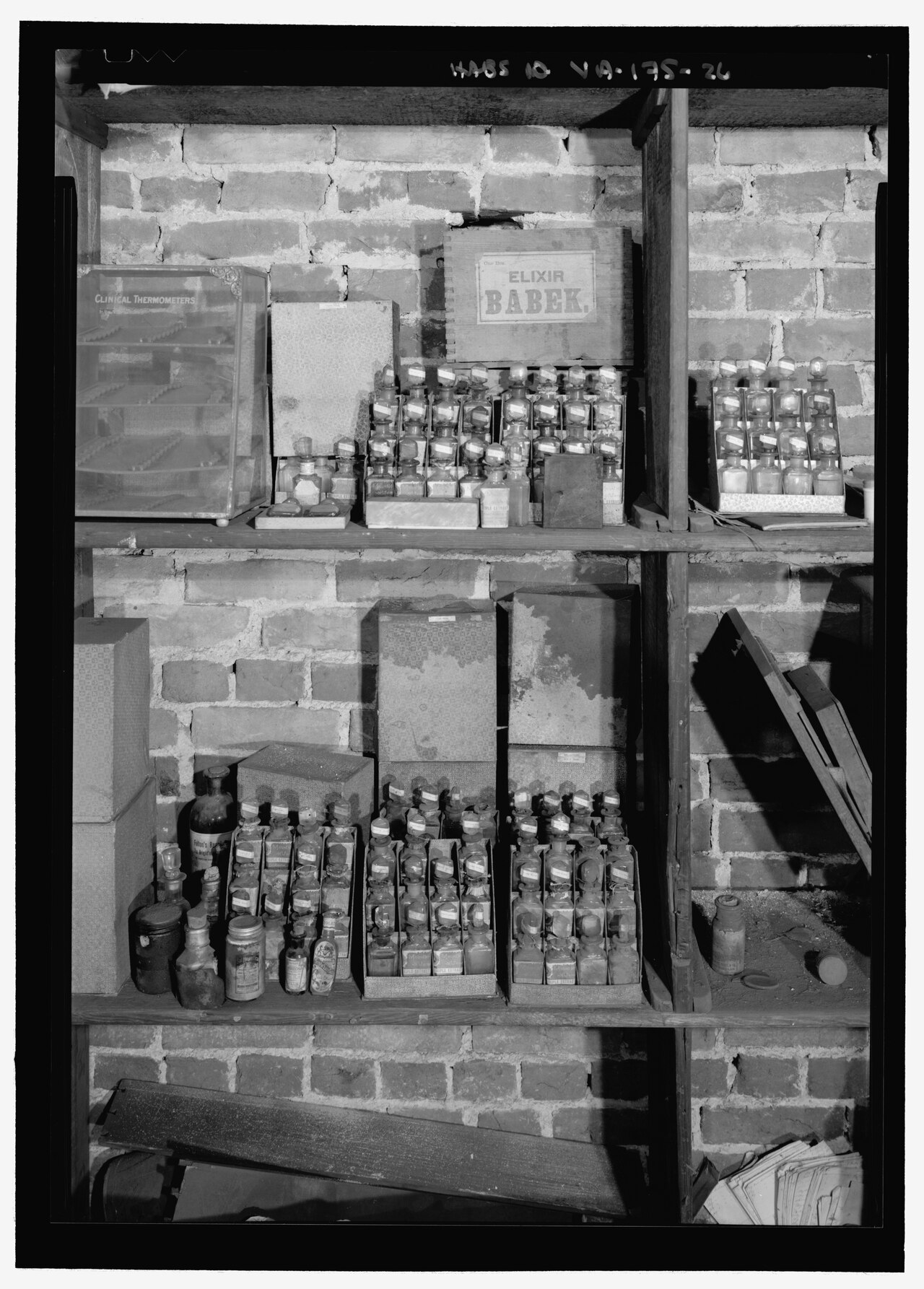 Black-and-white photograph of old apothecary shelves with elixir bottles, boxes and a thermometer case